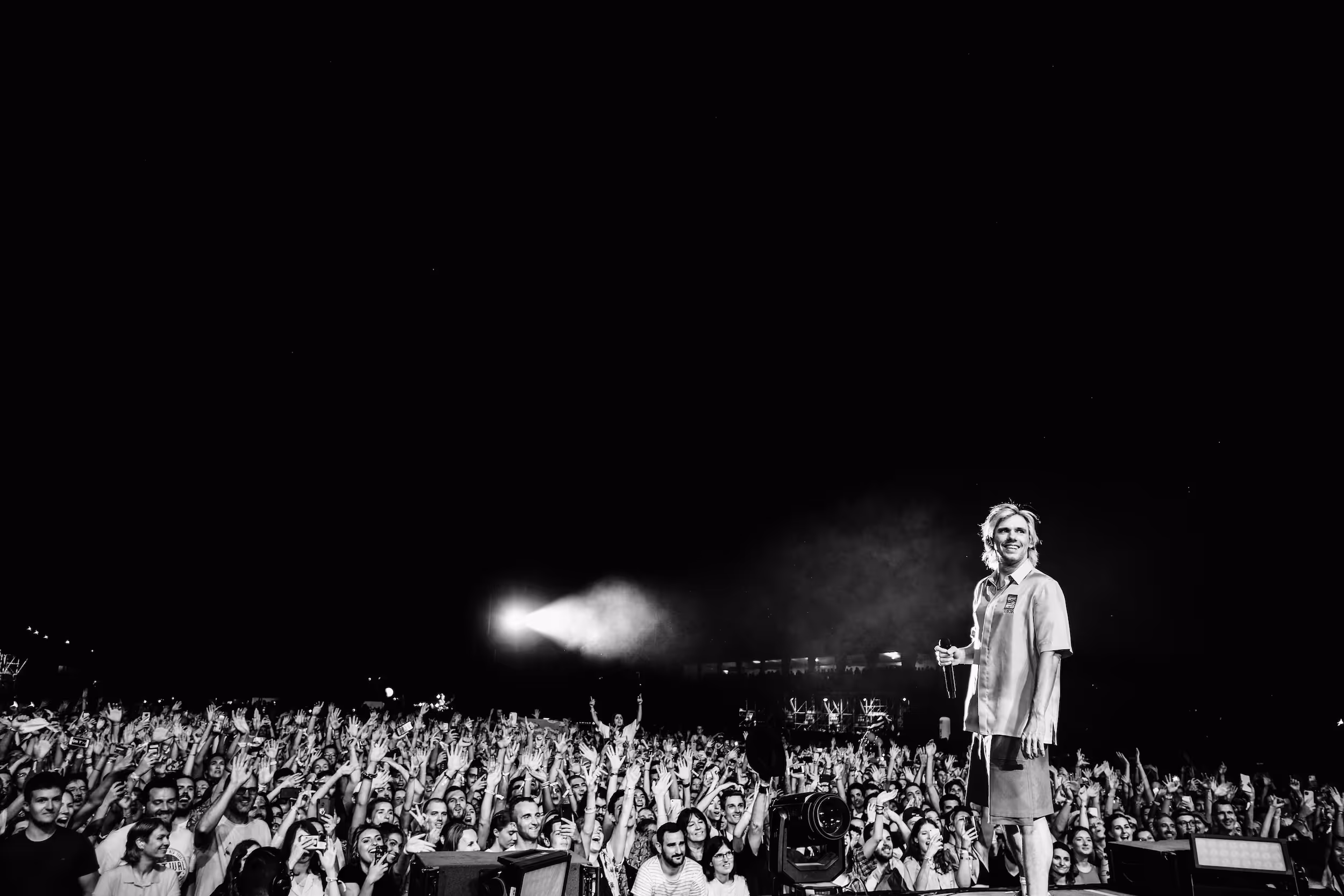 Black-and-white photo of a male performer holding a microphone and smiling on stage facing an enthusiastic crowd with raised hands at night.