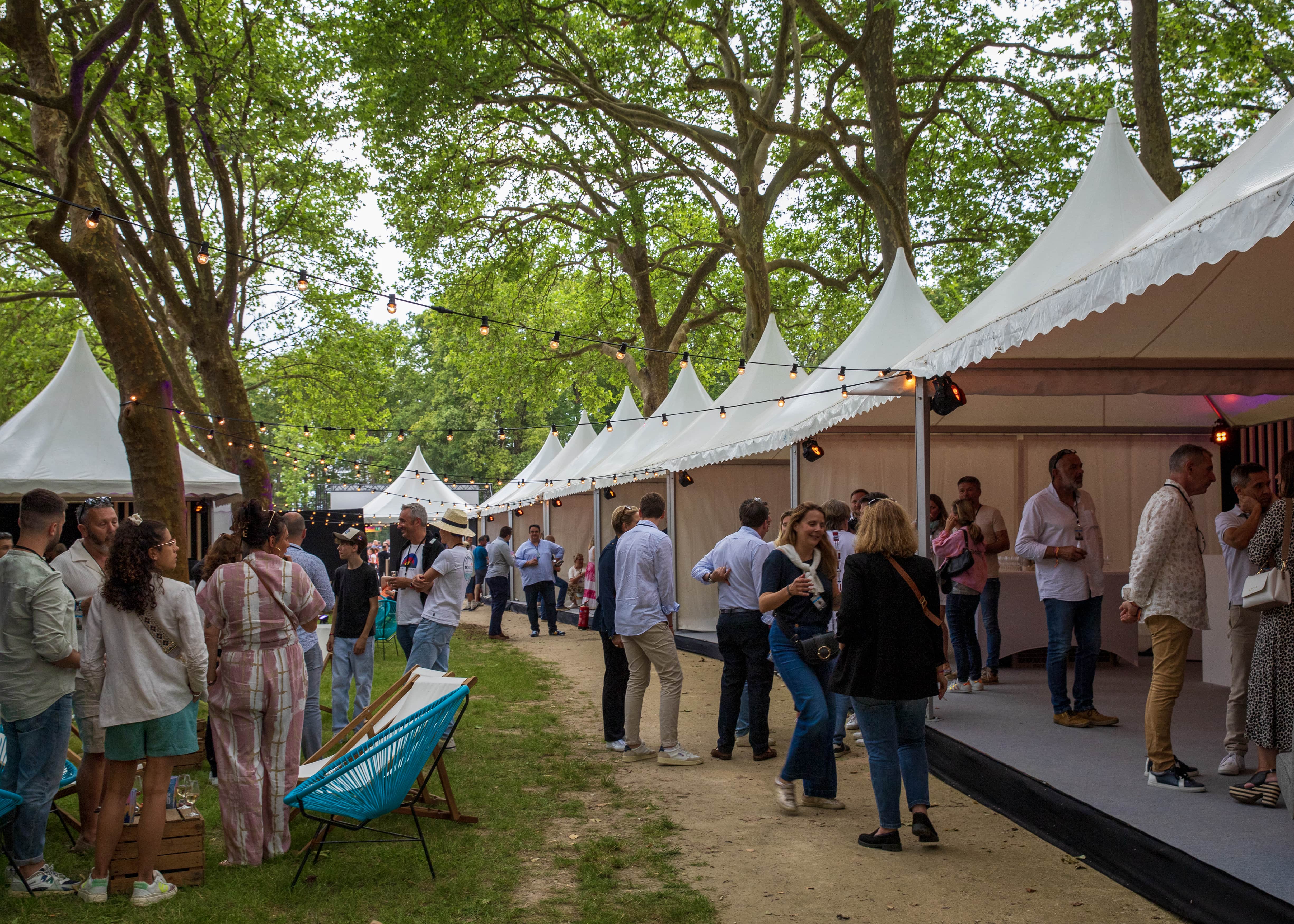 Outdoor event with people socializing near white canopy tents under string lights in a green park setting.