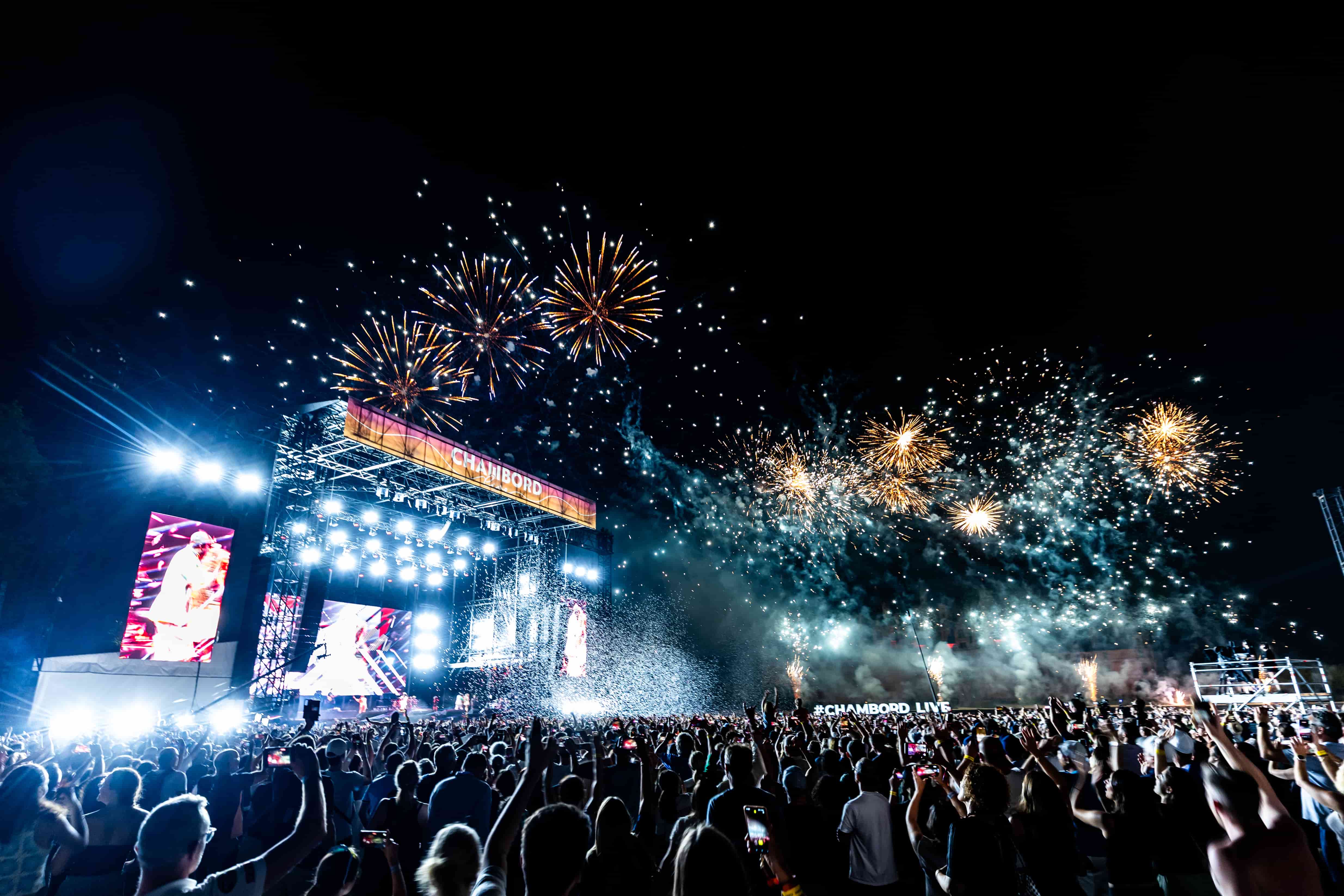 Crowd at a nighttime outdoor concert with a brightly lit stage, large screens, and fireworks lighting up the sky.