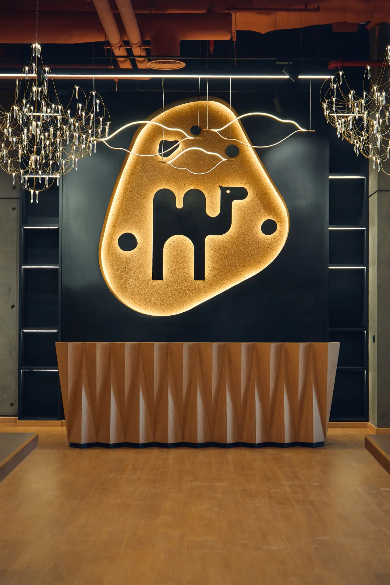 Reception area of Kamelizer Spaces Majarrah branch in Sheikh Zayed, featuring a modern interior with a stone wall, abstract camel logo installation, artistic hanging lights, and a golden reception desk.