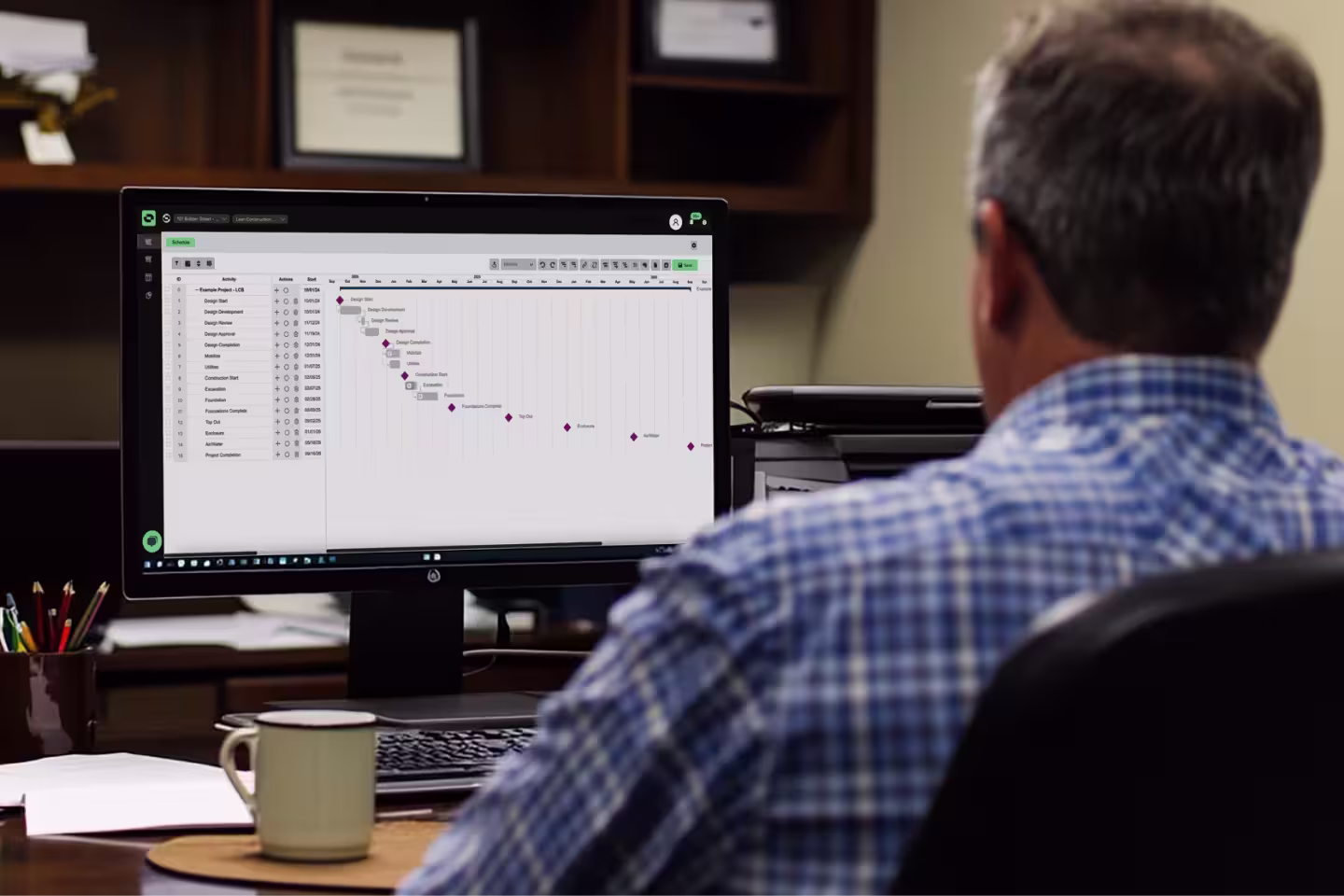 A scheduler sat at a desk in a construction office shown using the Outbuild web application on his laptop