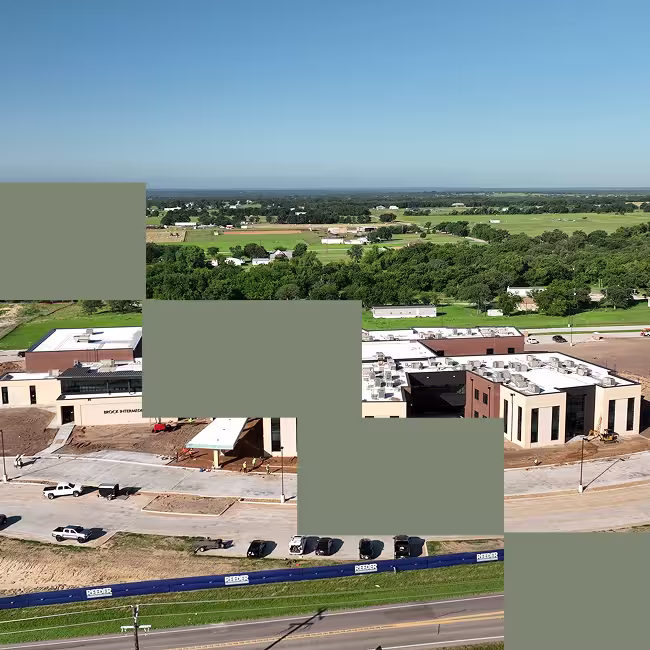 Aerial view of a large multi-building construction site near a road, with several parked vehicles and green fields in the background.