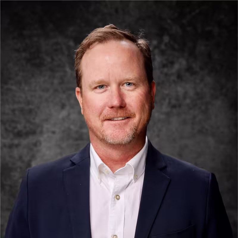 Man with light brown hair and beard wearing a navy blazer and white shirt against a dark background.