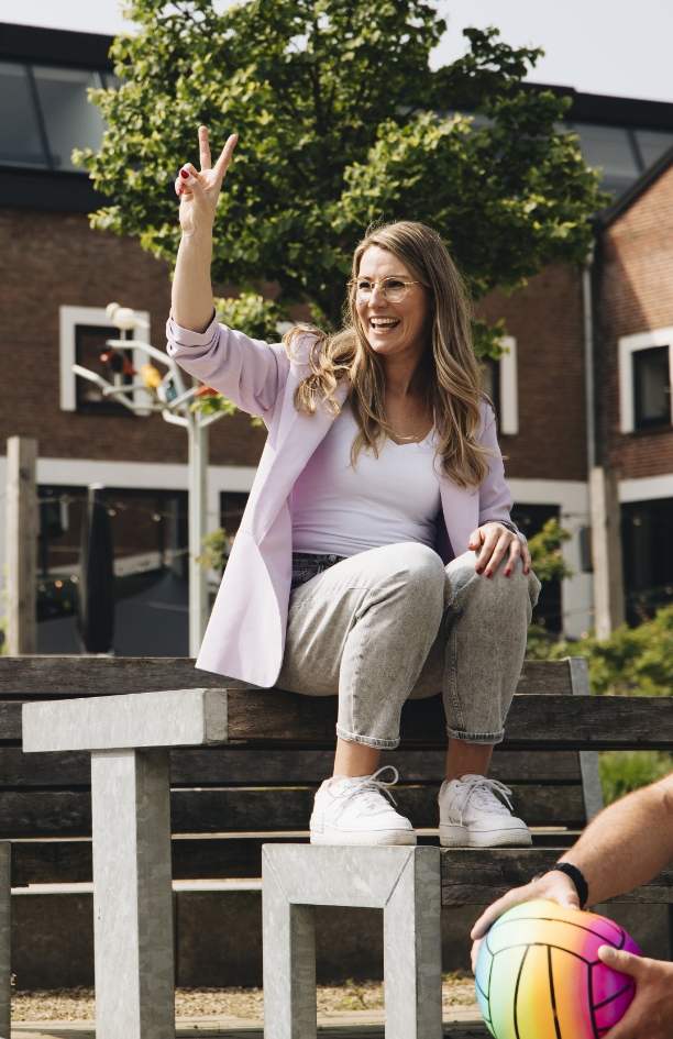 Trainer en coach Amanda Ronhaar van Toer de Werkvloer zit buiten of een tafel en steek twee vingers in de lucht.