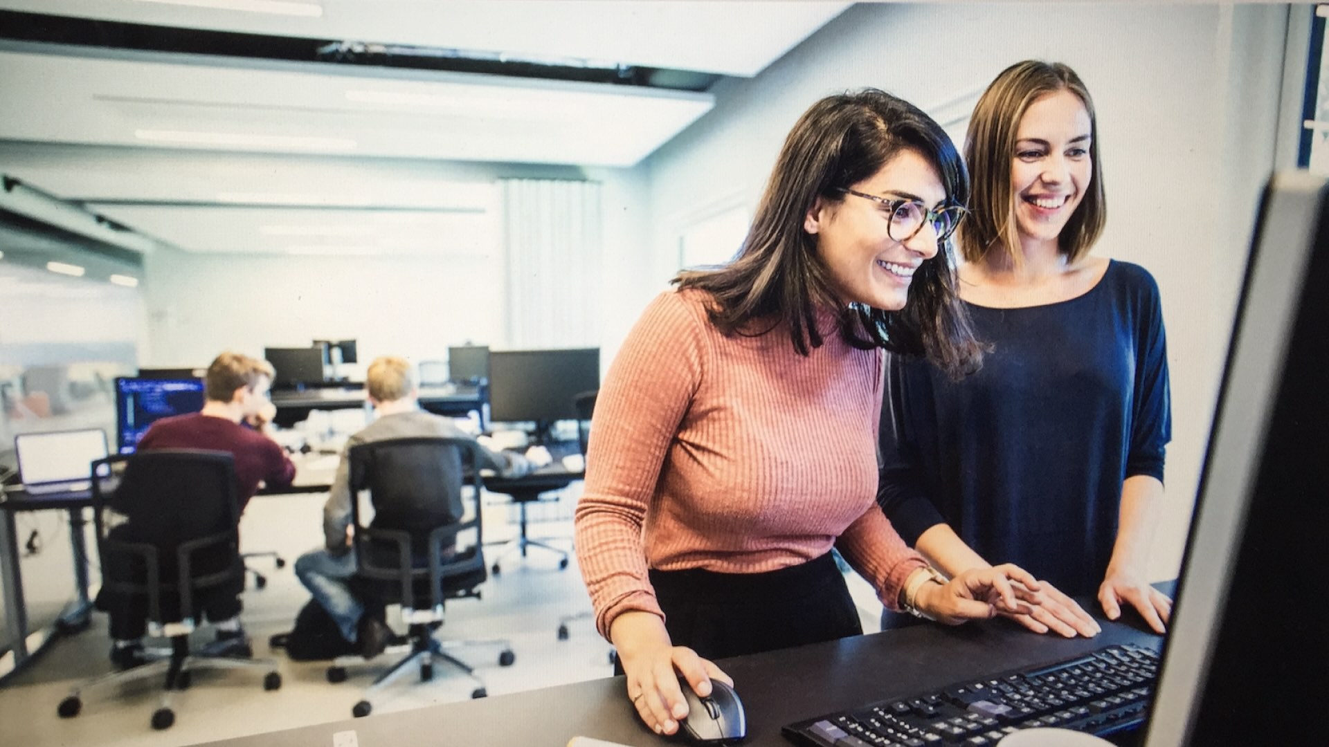Two people standing at a computer and smiling. In the background, two other people are seated at desks with multiple monitors.