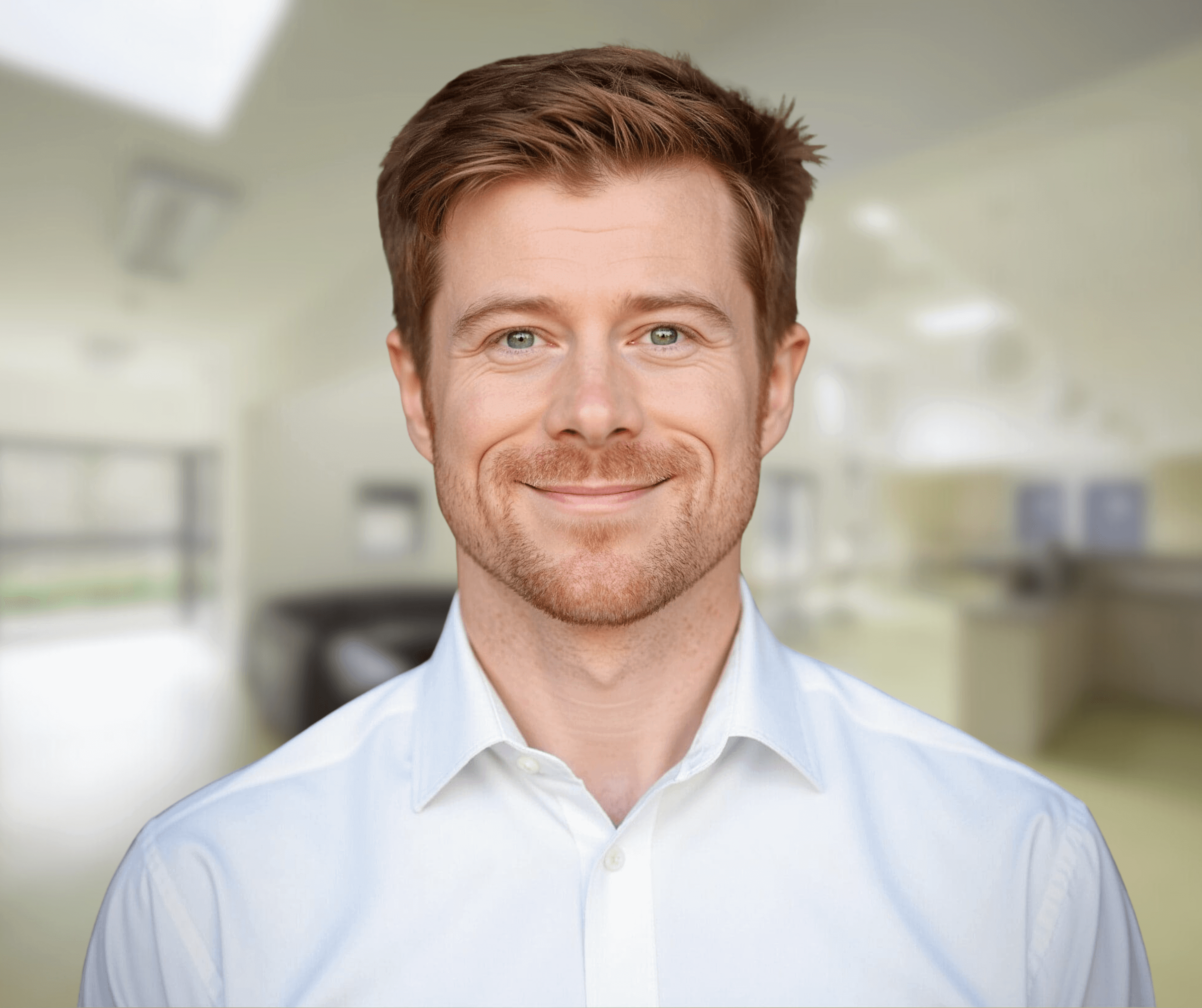 A smiling man with short light brown hair and a trimmed beard, wearing a white button-up shirt, standing indoors in a softly lit modern room with blurred furniture and windows in the background.