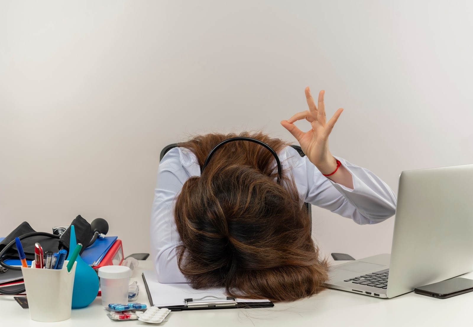 Eine Frau signalisiert per Hand "Alles okay!", hat jedoch ihren Kopf auf der Bürotischplatte abgelegt.