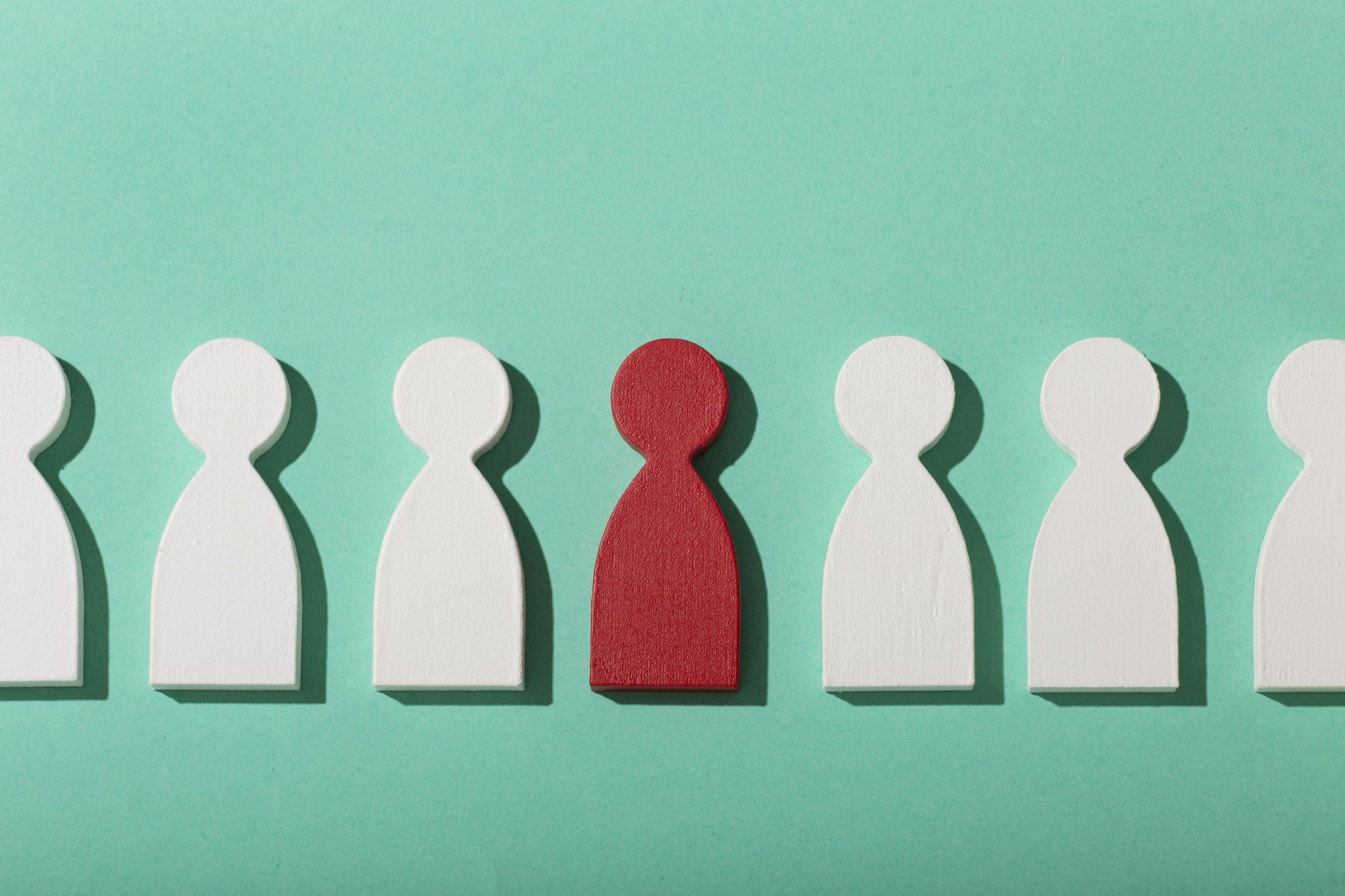 Eine rote Figur steht allein zwischen weißen Figuren und symbolisiert den Individualist beim Imposter-Syndrom. Das Bild zeigt das Gefühl, anders zu sein, Hilfe von Mitmenschen schwer anzunehmen und den eigenen Erfolg allein beweisen zu müssen.