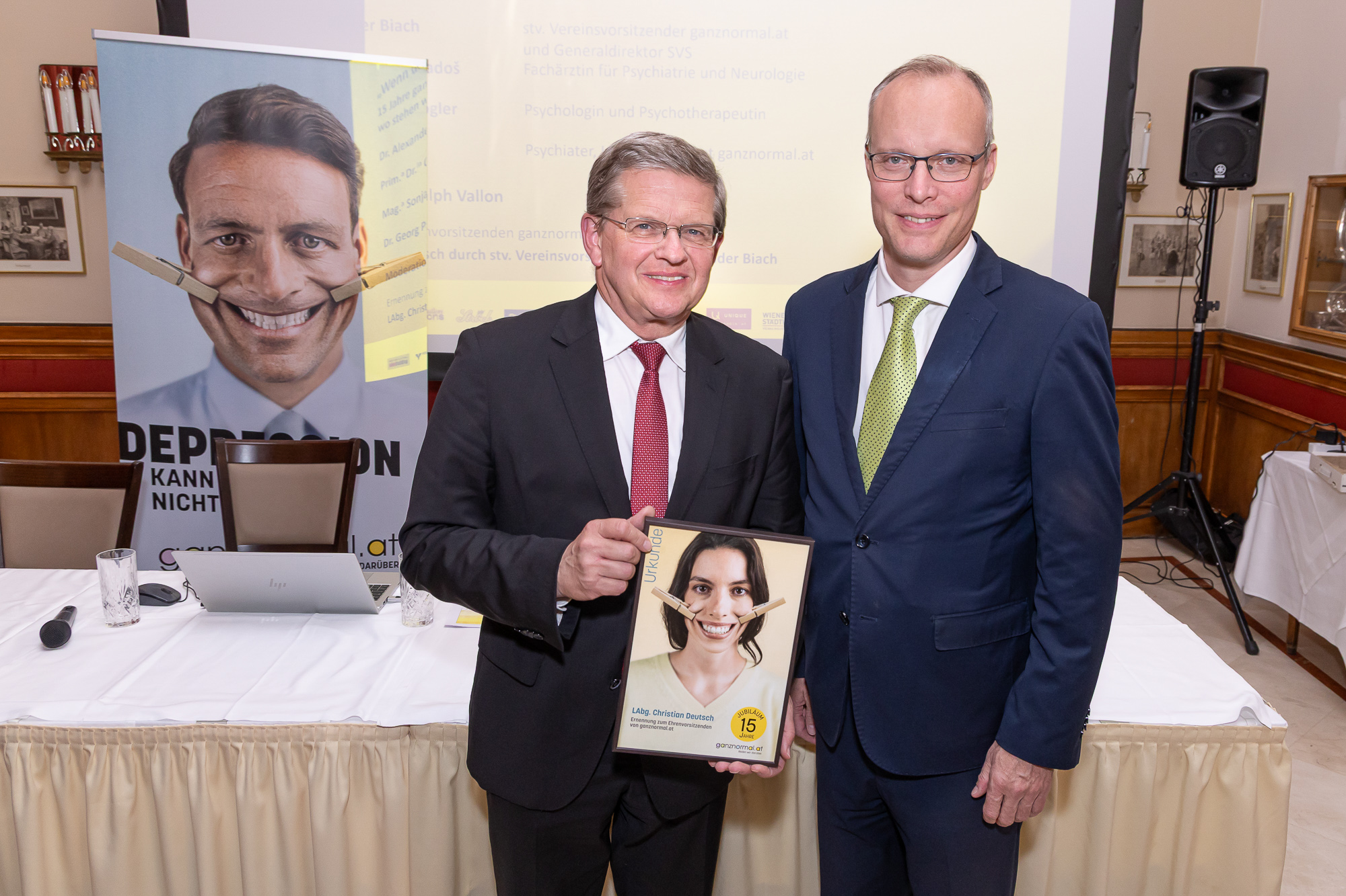 Zwei Männer im Anzug präsentieren eine gerahmte Auszeichnung vor einem Kampagnenplakat von ganznormal.at.