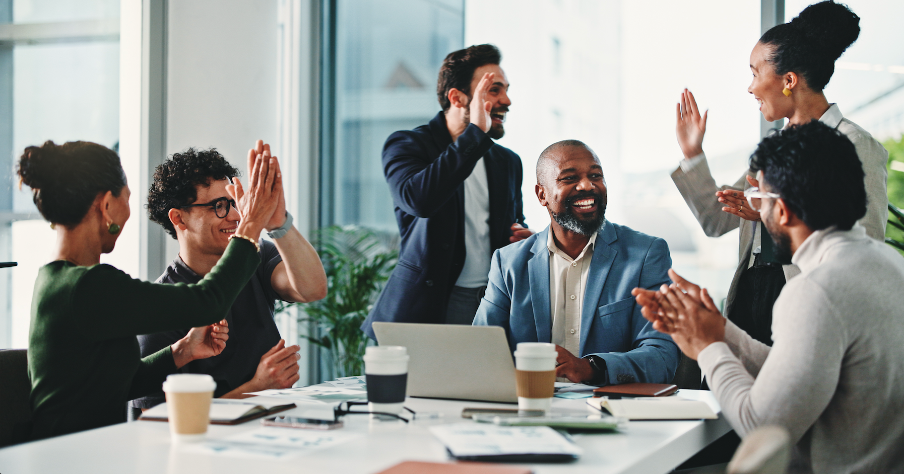 A diverse team of professionals celebrating around a meeting room table, high-fiving and applauding — illustrating the moment of business success that can precede a sale or exit.