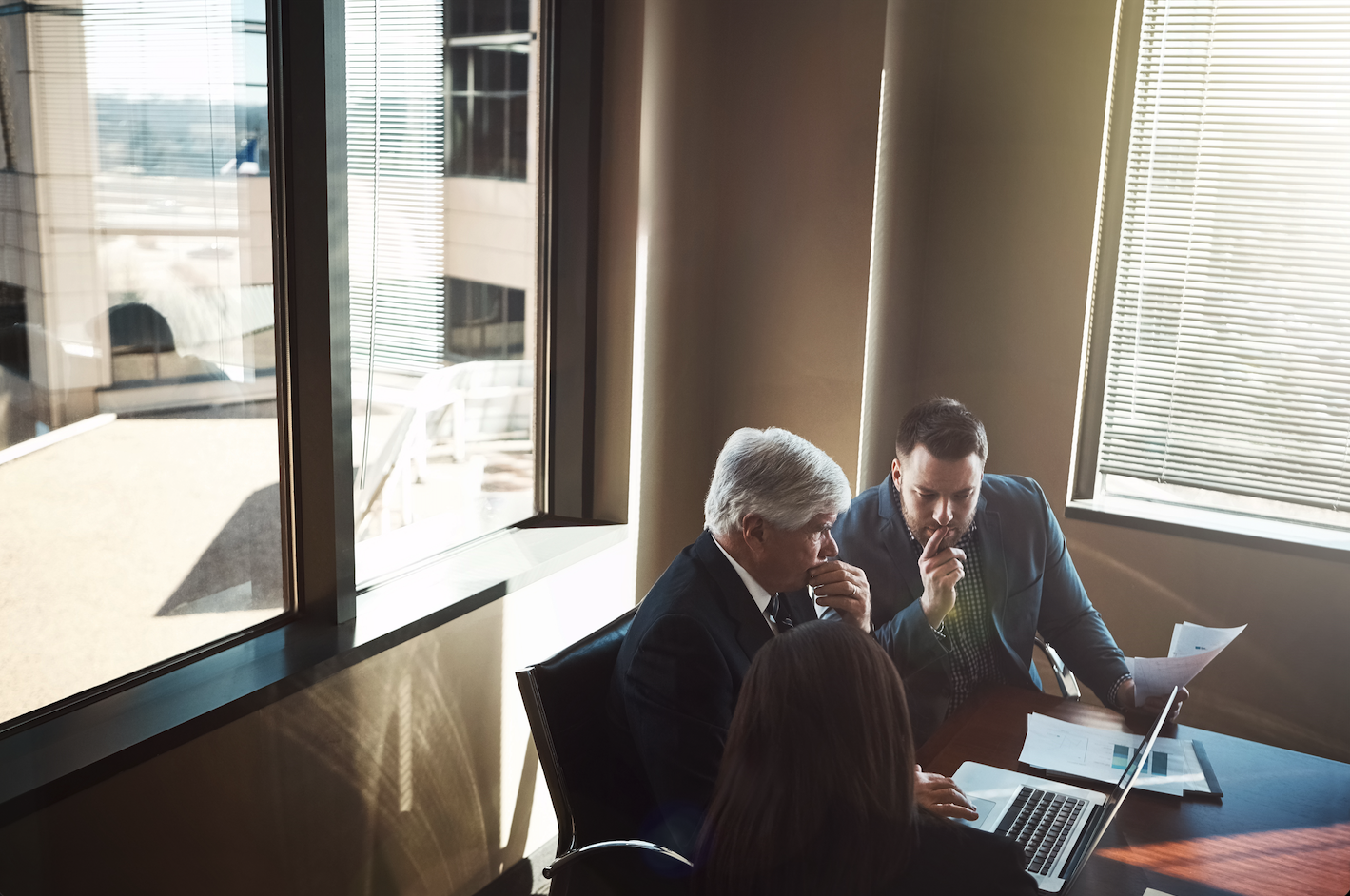 Three professionals in serious discussion around a laptop and documents in a boardroom — illustrating the shift from operational business risk to the considered, adviser-led approach to investment risk that follows a company sale.
