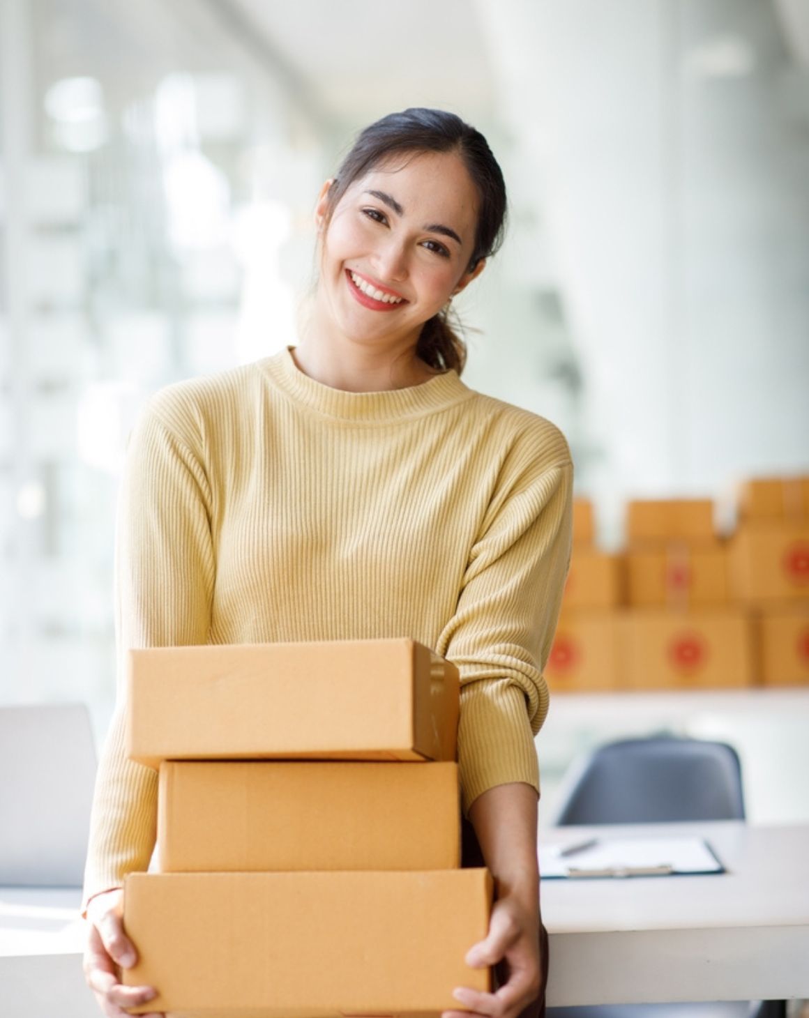 a woman stands in an office, balancing a stack of boxes in her arms