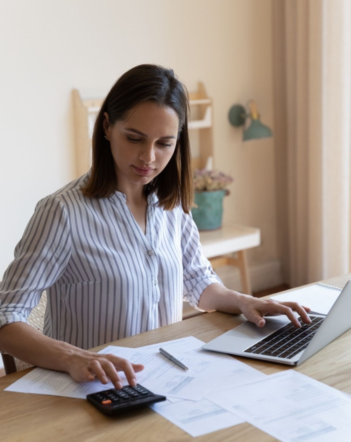 a woman sits at a table, focused on her laptop with calculator