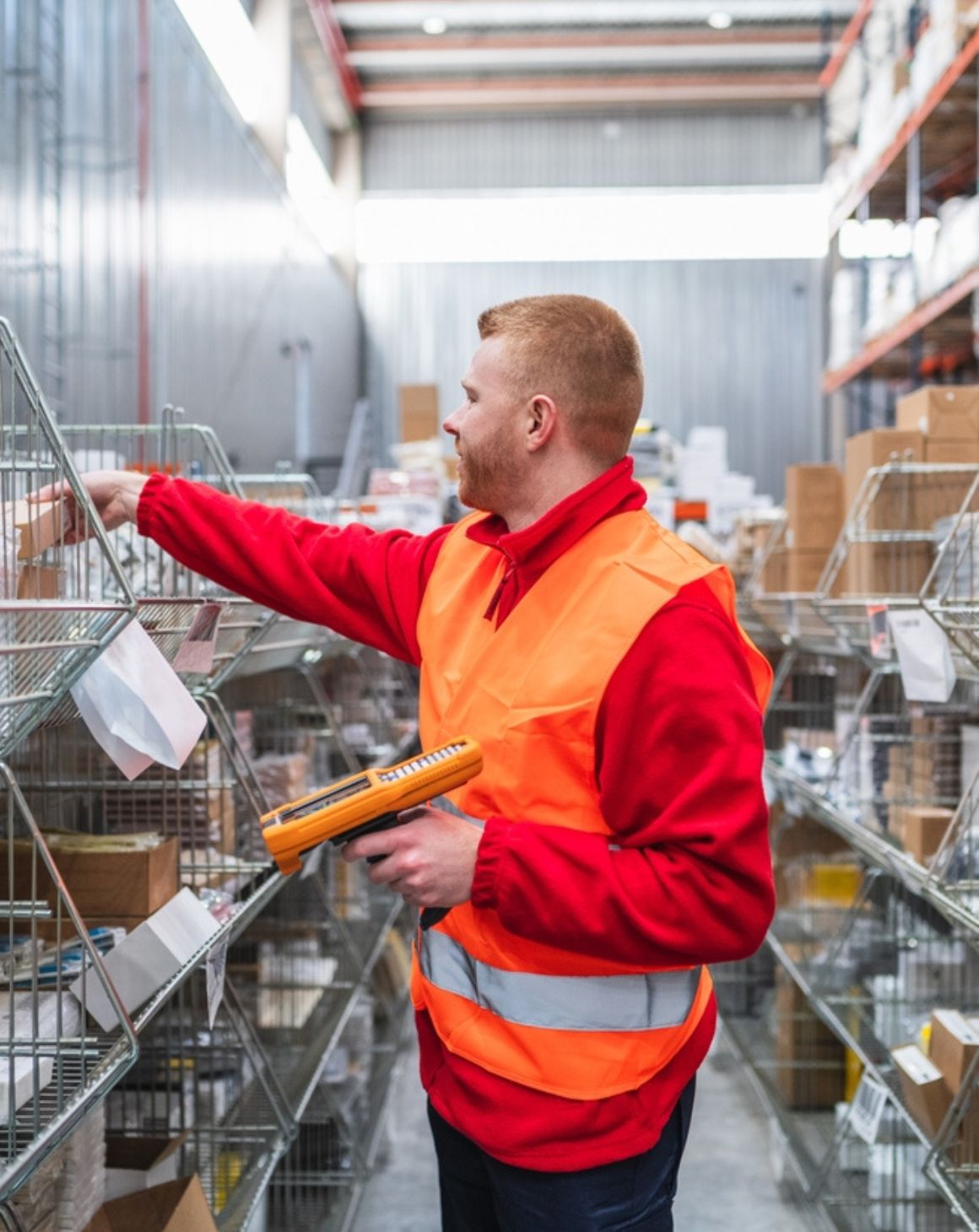 a man in a warehouse wears an orange vest and holds a clipboard
