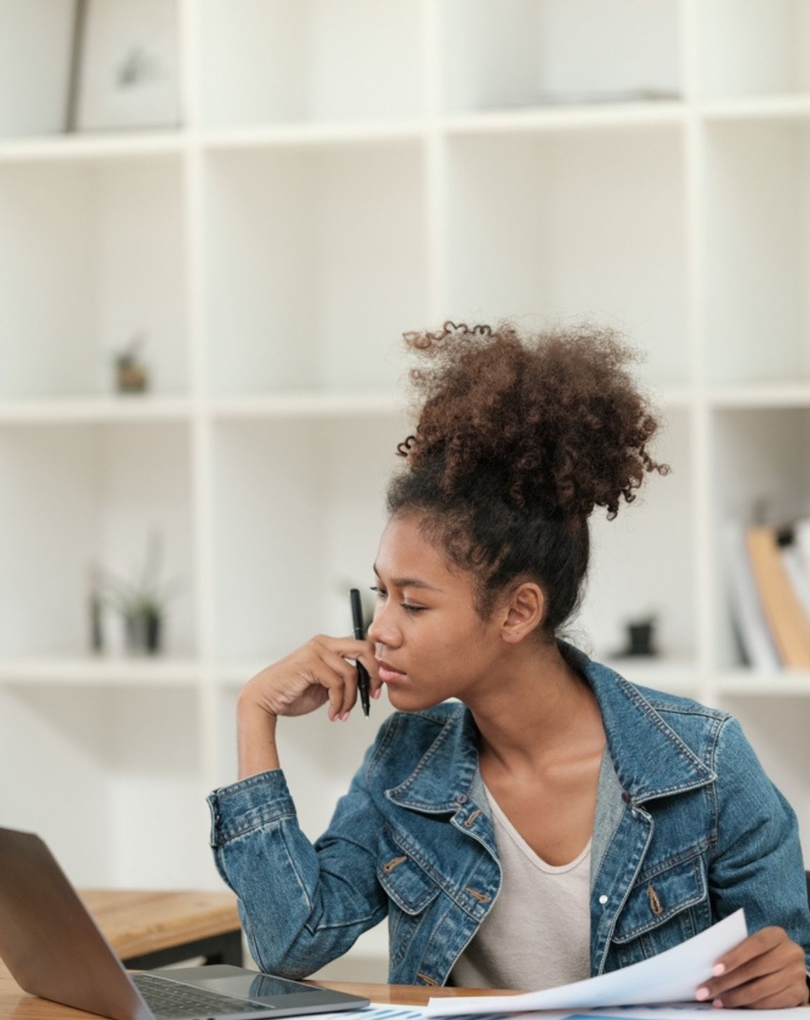 woman sits at desk in office setting looking at laptop with documents in hand