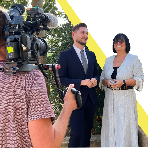 A cameraman filming a smiling man in a suit and woman in a white dress with sunglasses, outdoors near trees.
