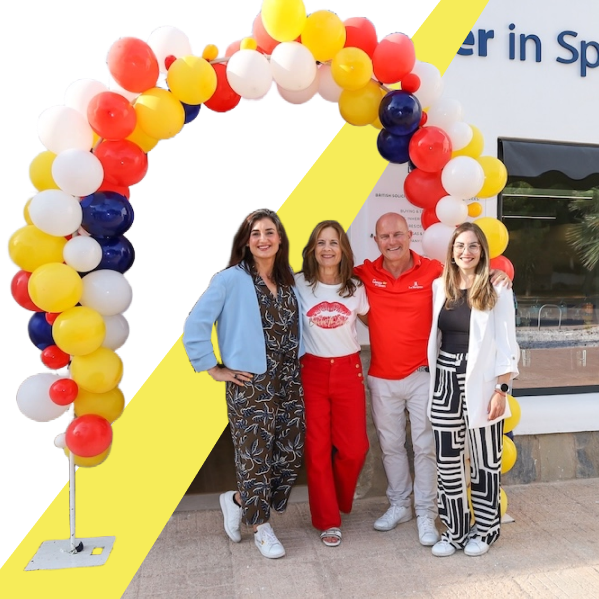 Four smiling adults standing under a colorful balloon arch with red, yellow, white, and blue balloons outside a building.