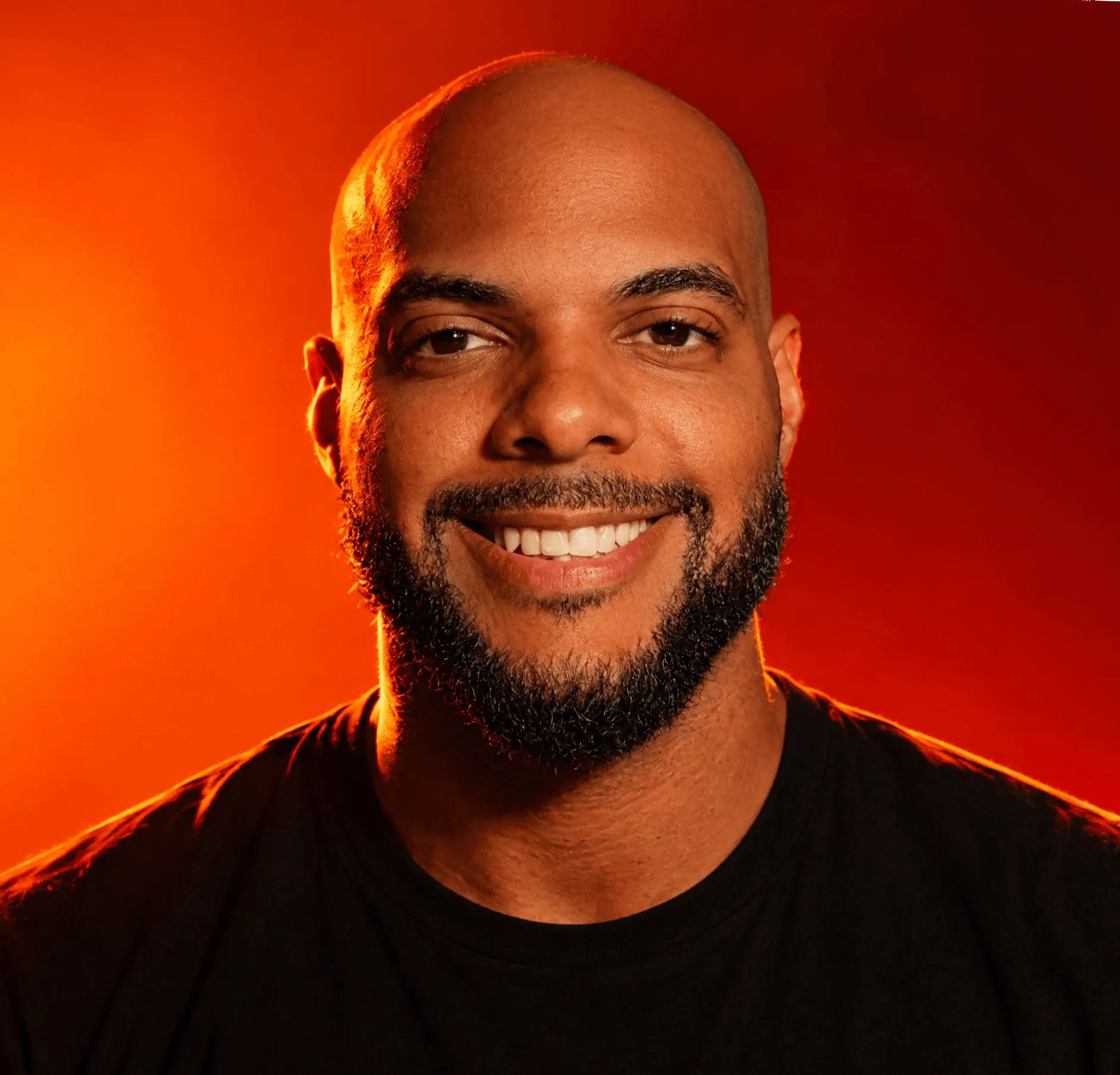 Portrait of a bald man with a beard wearing a black shirt against a dark background.