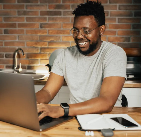 Smiling man wearing glasses and a gray t-shirt working on a laptop at a wooden table with a notebook and smartphone.