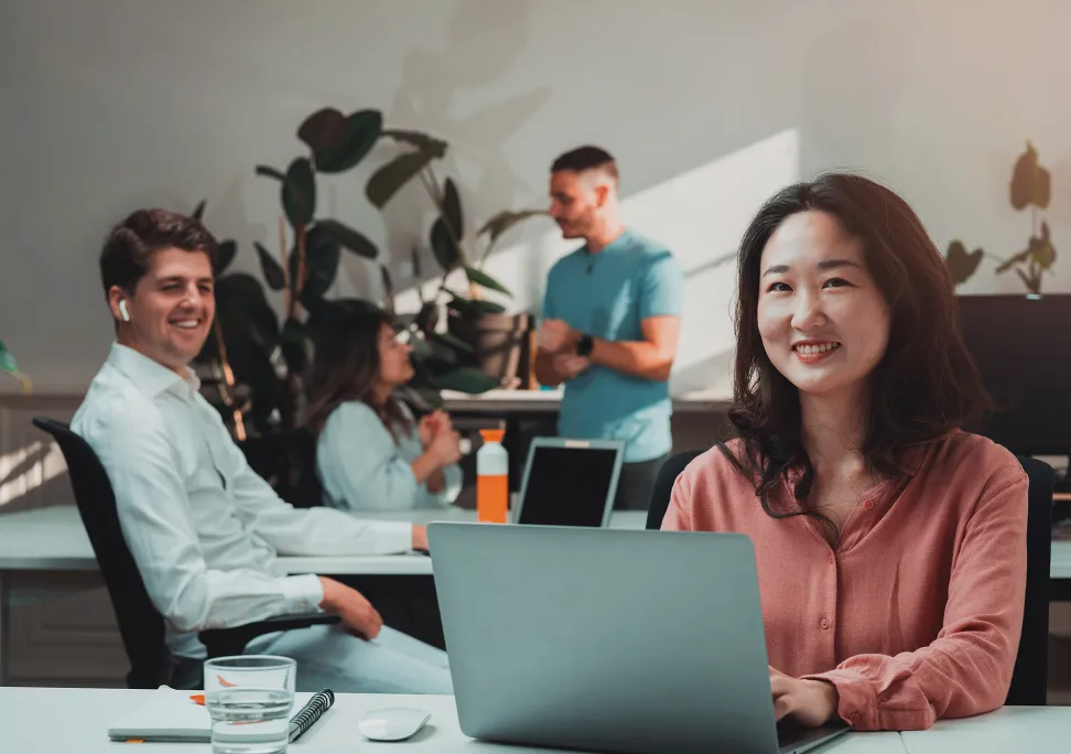 Smiling young woman working on a laptop at a desk with colleagues discussing in the background in a bright office.