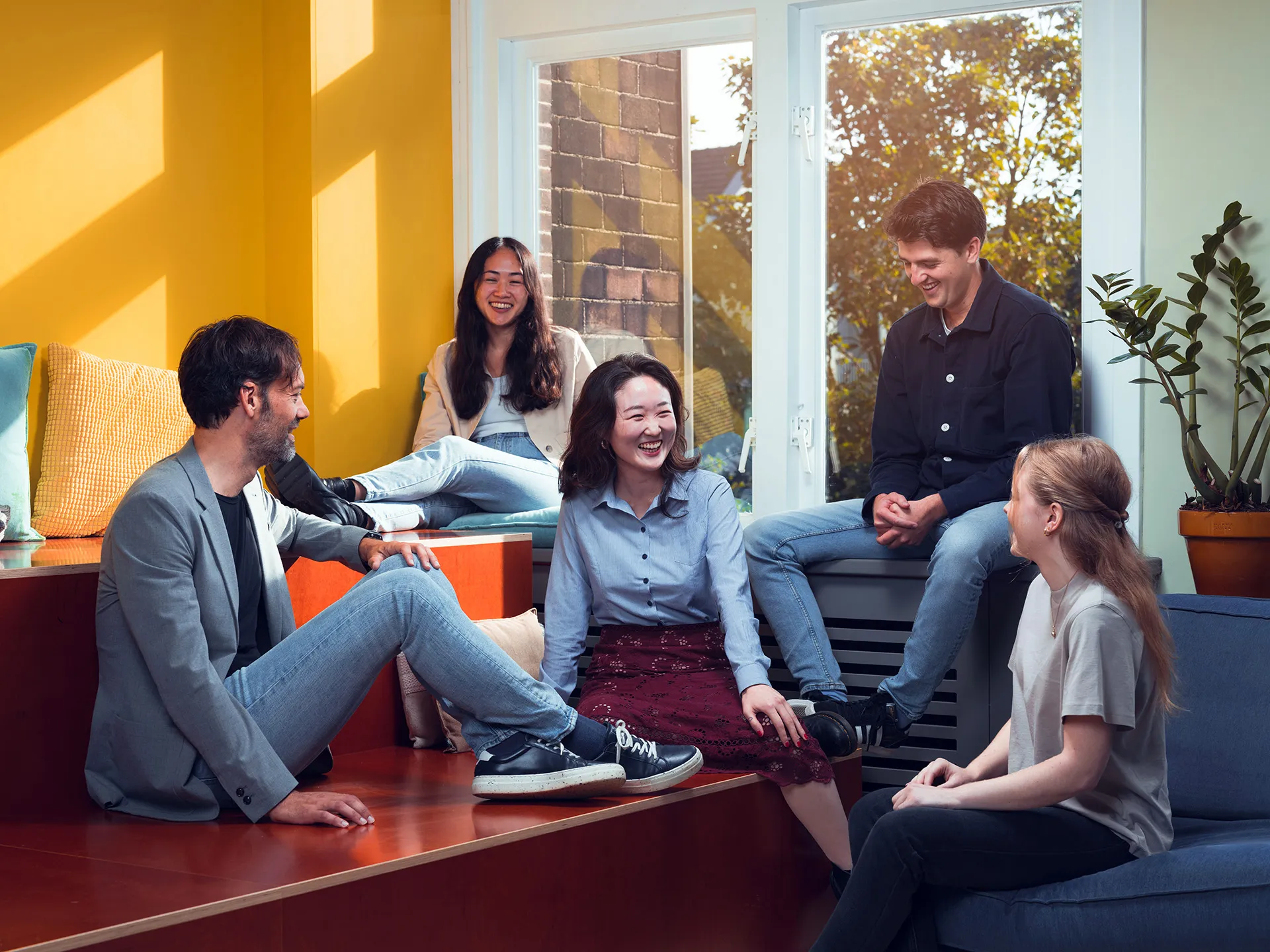 Five young adults laughing and chatting in a bright room with yellow walls and large windows.