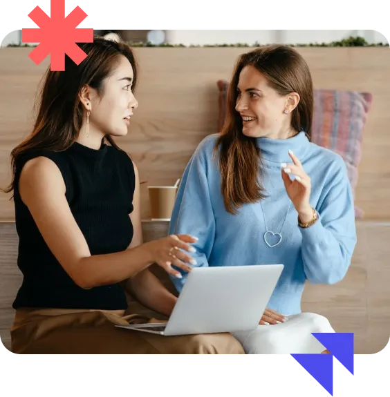Two women sitting and smiling at each other while using a laptop, with colorful geometric shapes in the corners.