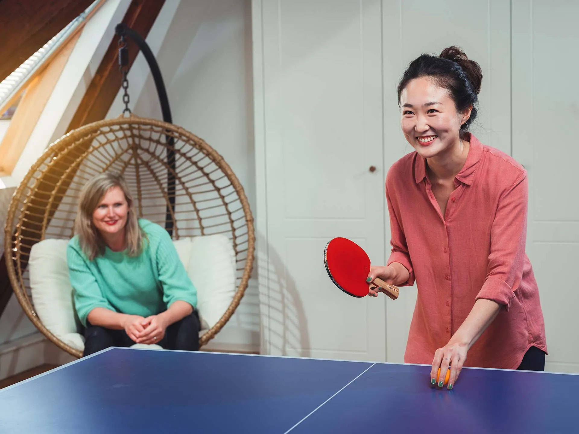 Smiling woman in a pink shirt holding a table tennis paddle and ball, with another woman sitting in a hanging chair in the background.