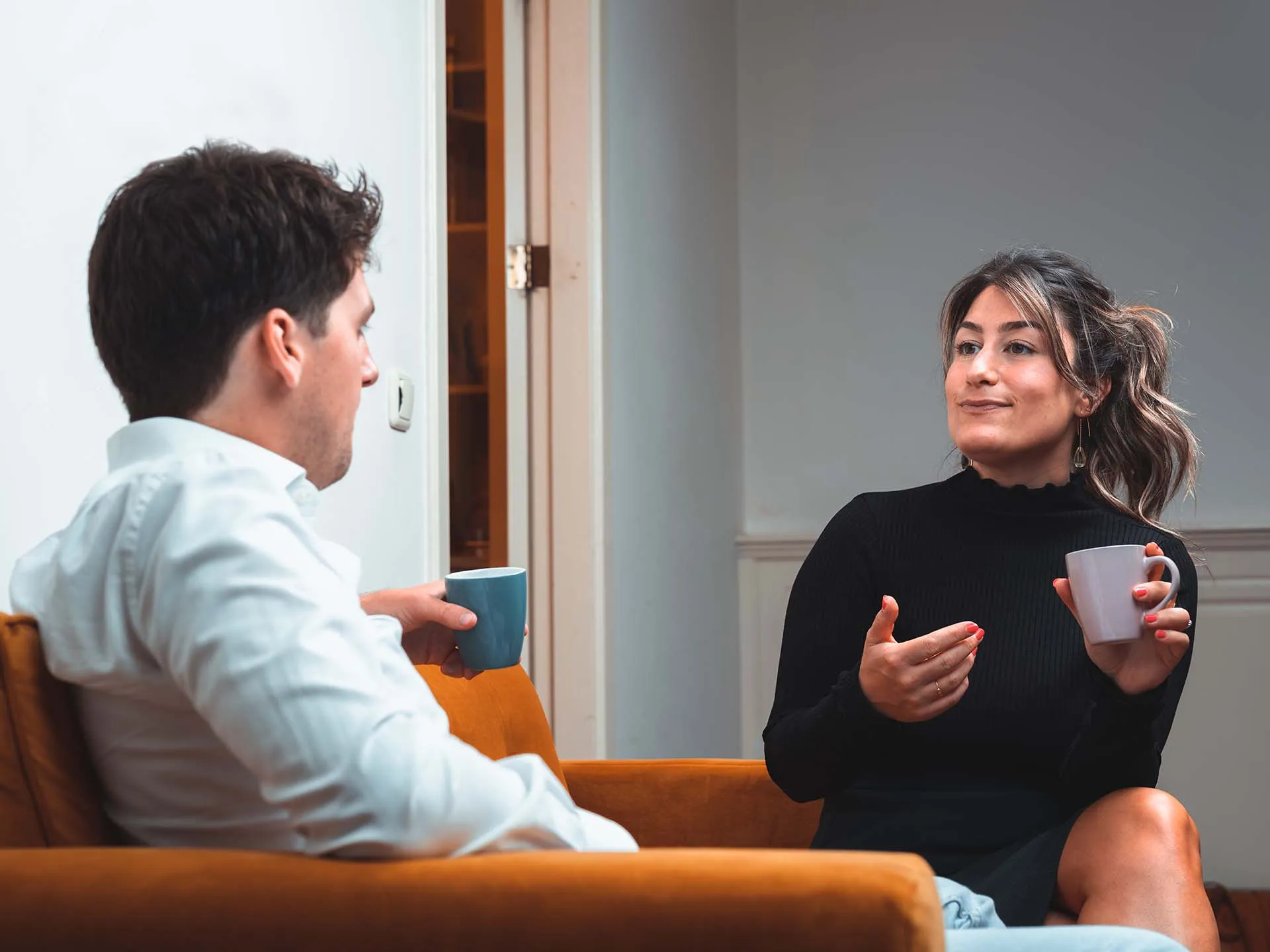 Two people sitting on a brown couch drinking coffee, engaged in a conversation in a cozy room.