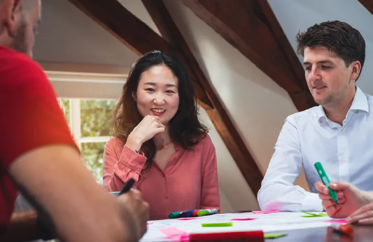 Three colleagues in a meeting room with wooden beams, discussing and using colorful markers on paper.