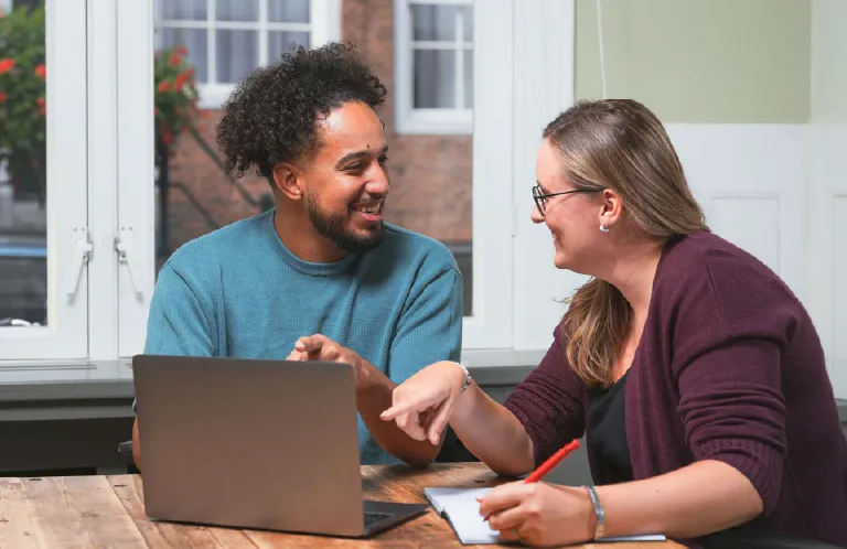 Two colleagues smiling and discussing work over a laptop at a wooden table near a window.