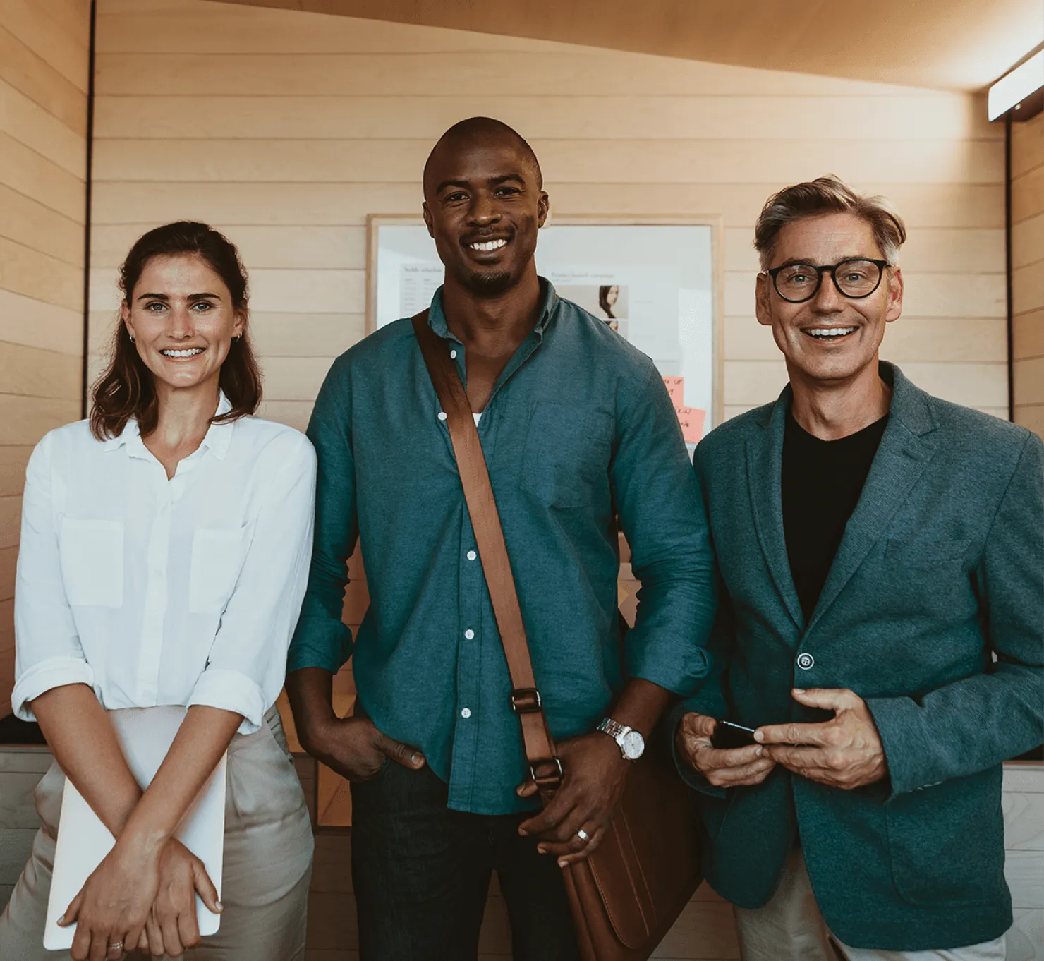 Three professionals standing indoors: a woman holding a laptop, a man with a shoulder bag, and a man wearing glasses and a blazer, all smiling at the camera.