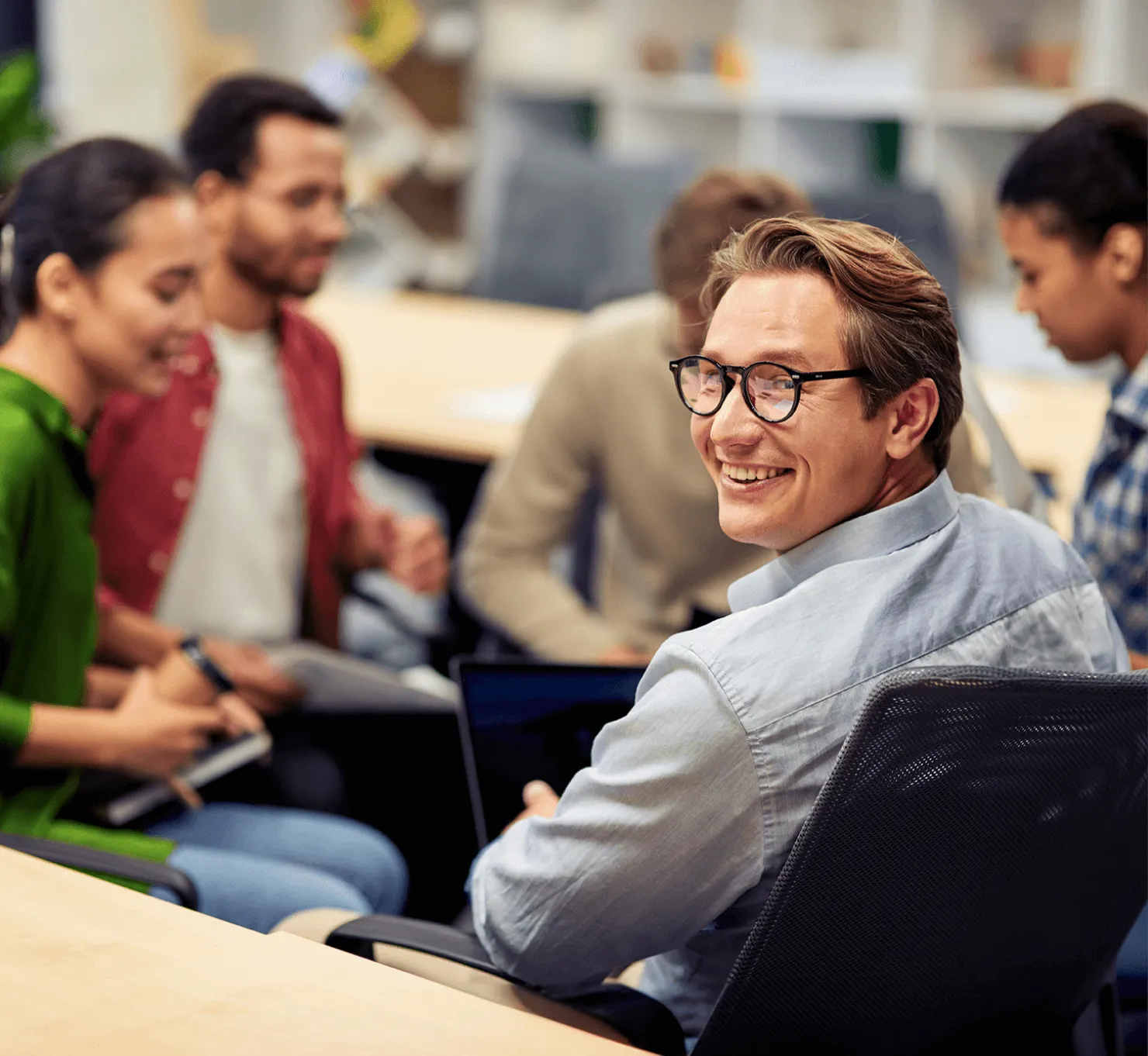 Smiling man with glasses sitting and working on a laptop in a group meeting with four other people.