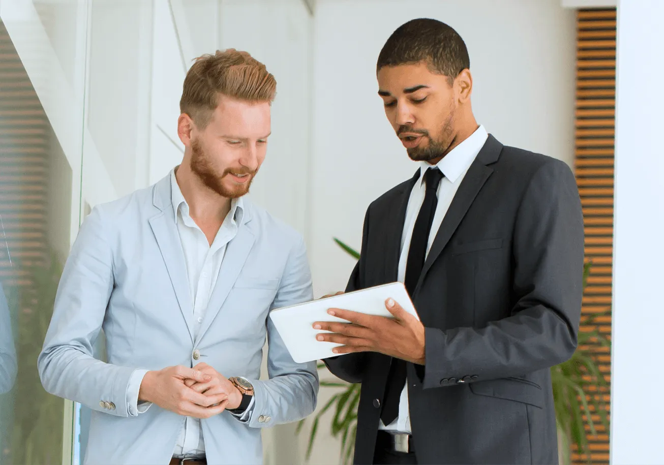 Two businessmen in suits discussing information on a digital tablet in an office.