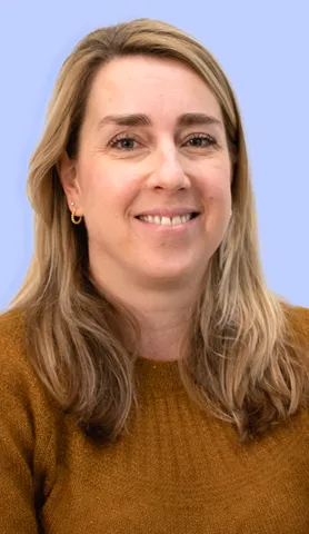 Smiling woman with long blond hair wearing a brown sweater and gold hoop earrings against a light blue background.