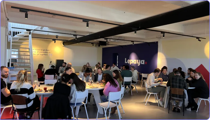 Group of people sitting around tables in a modern office cafeteria with Lepaya logo on a navy blue wall.