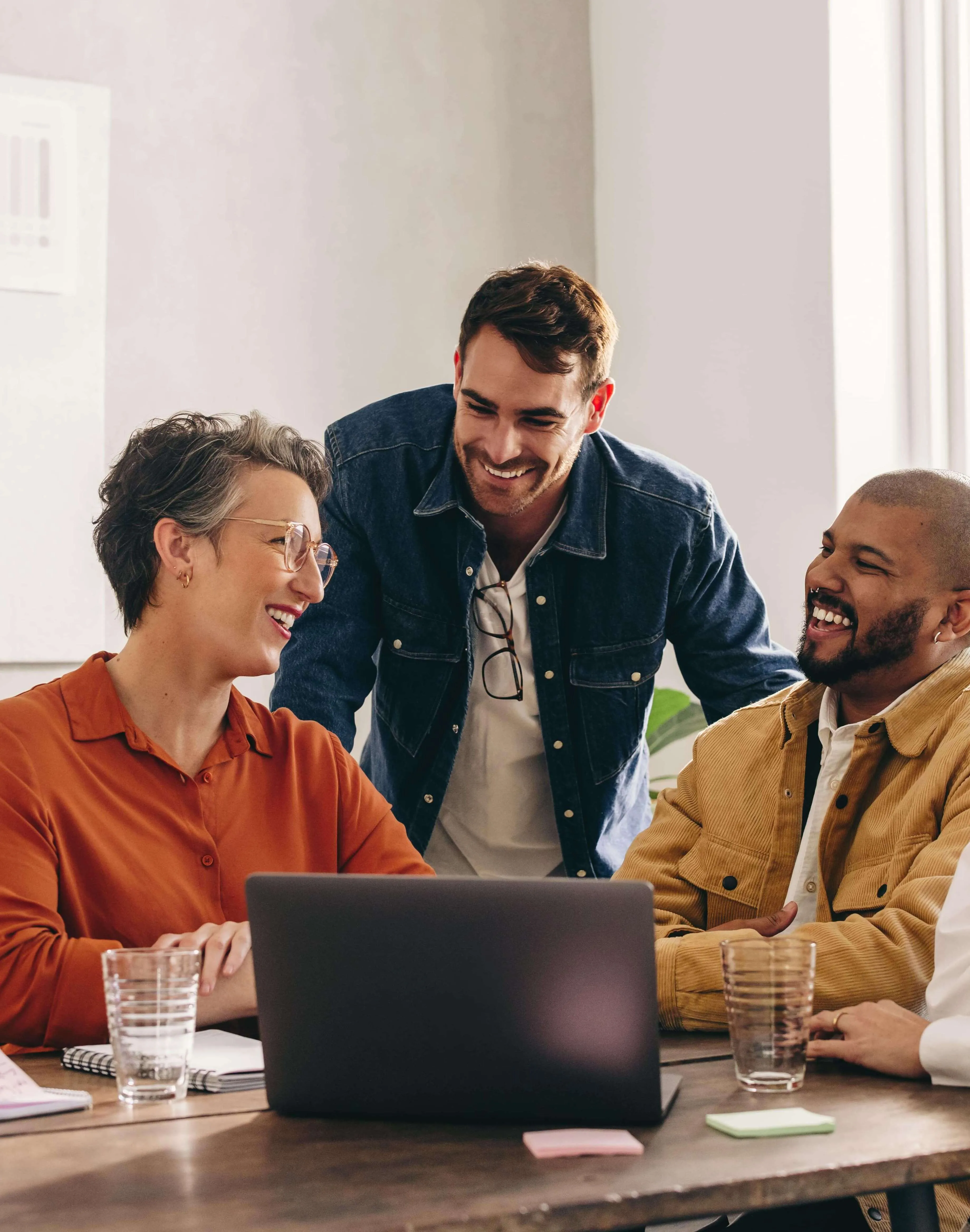 Three colleagues smiling and discussing work around a laptop in a bright office.