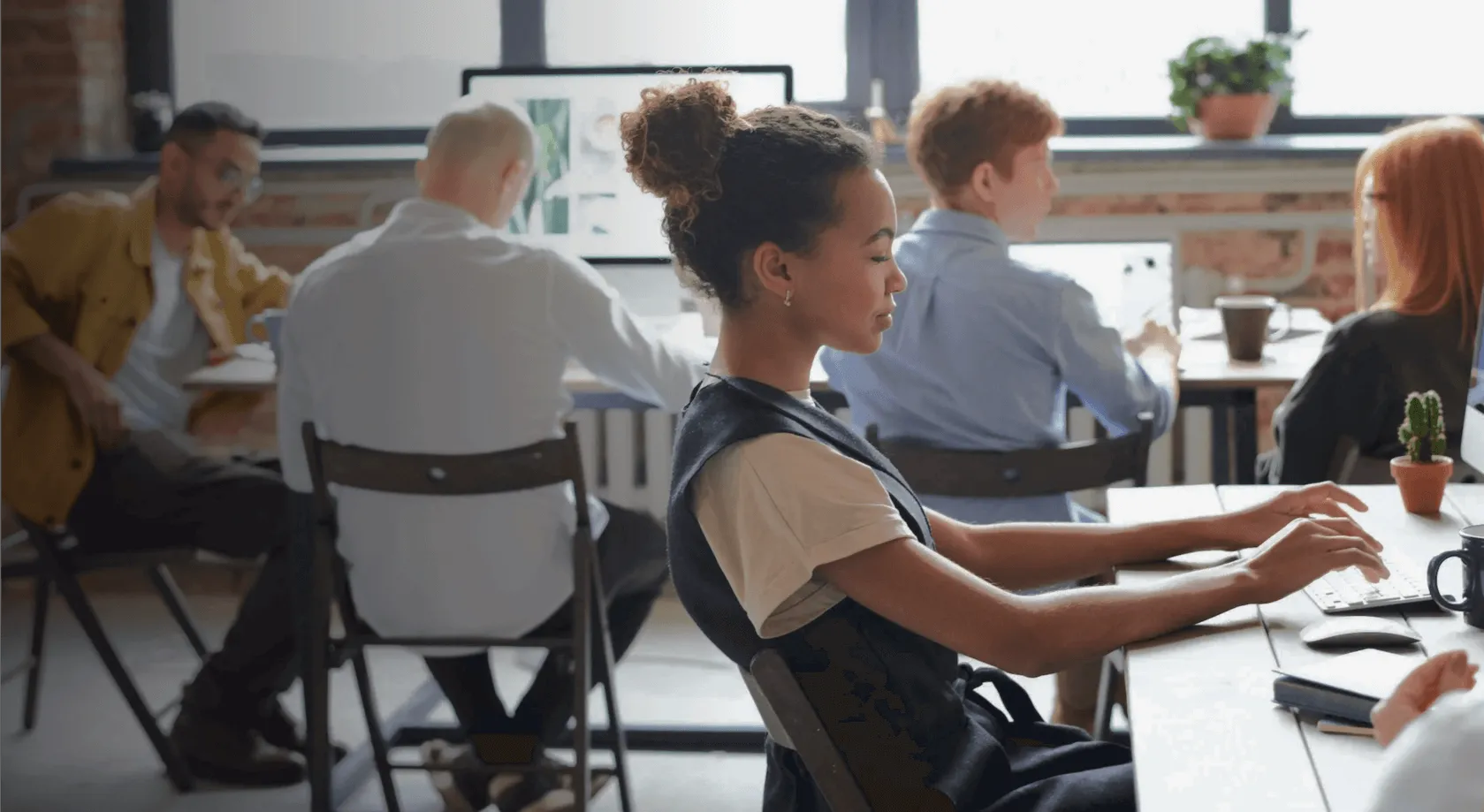 Young woman typing on a keyboard in a modern office with colleagues working at desks around her.