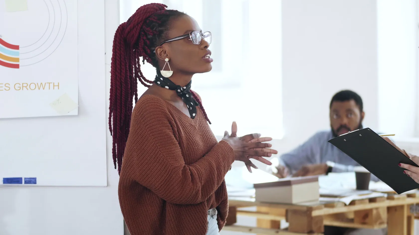 Woman with red braided hair and glasses speaking in an office with a whiteboard showing sales growth and colleagues working in the background.