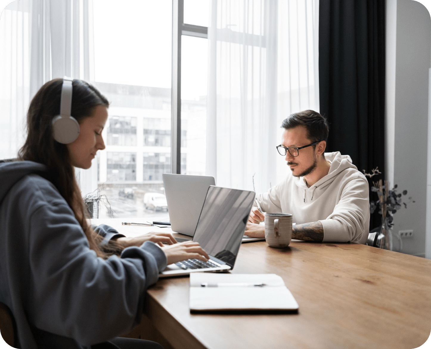 Two people working on laptops at a wooden table by a window with sheer curtains, one wearing headphones and the other writing with a mug beside him.