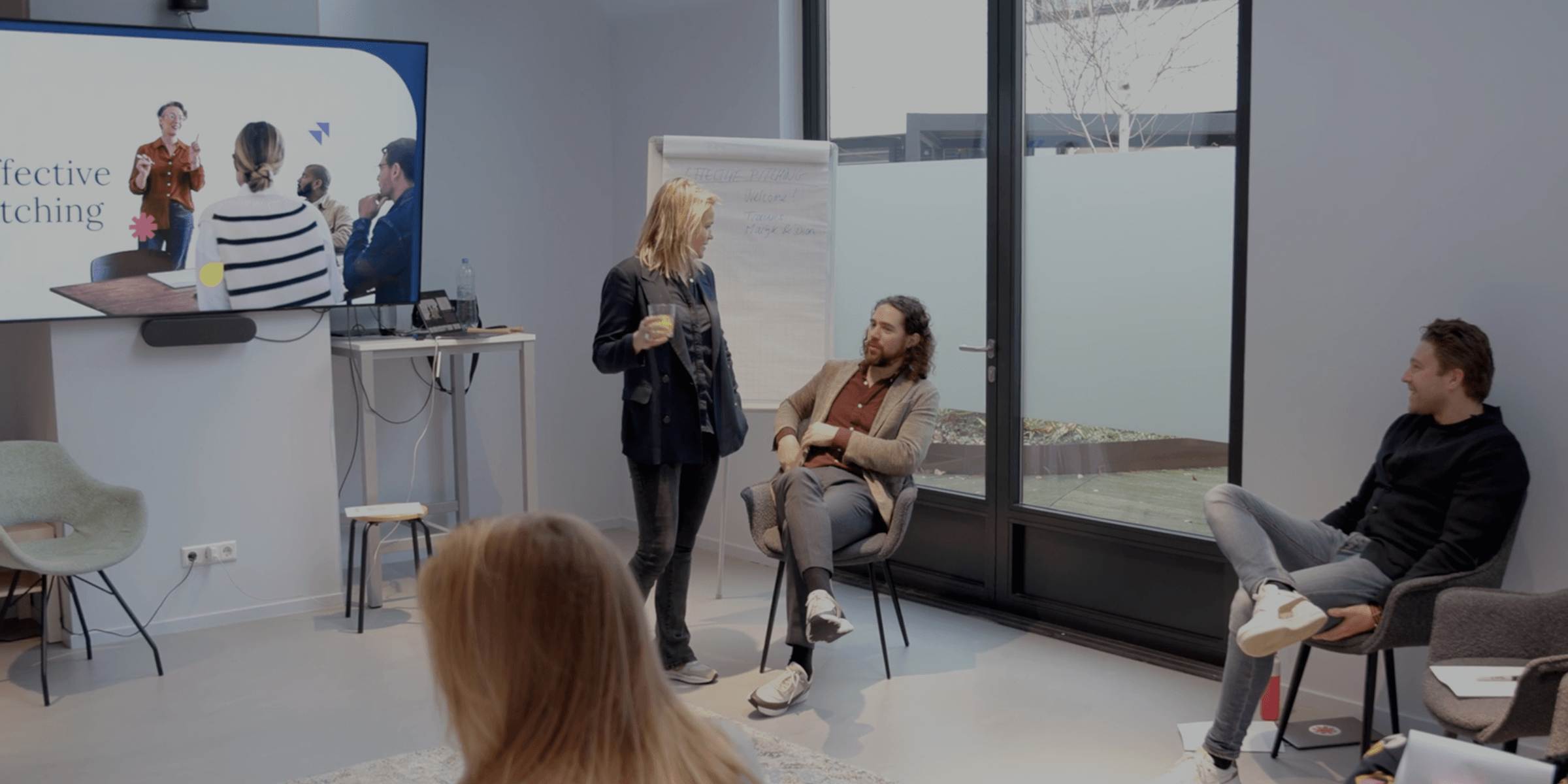 A small group of people in a modern meeting room, with one woman standing and holding a drink, two men seated and chatting, and a screen displaying a presentation on effective pitching.
