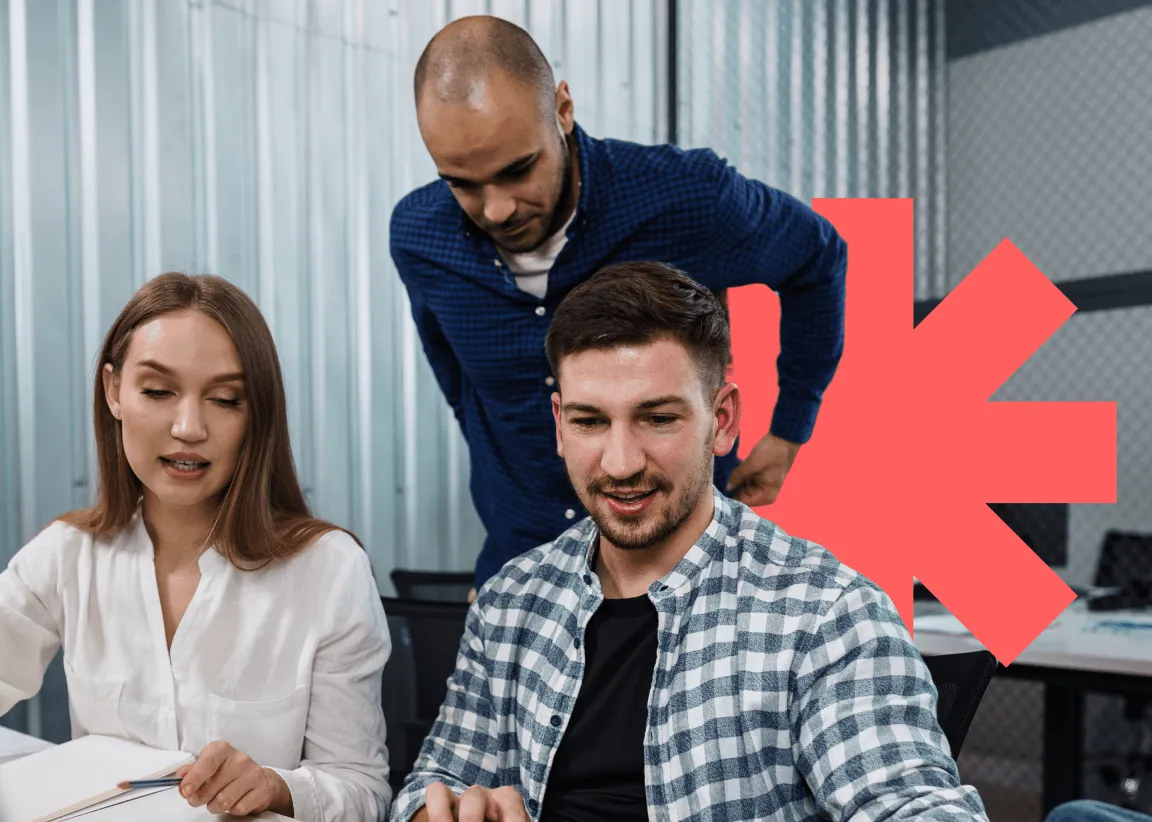 Three colleagues working together in an office with a man standing and two seated discussing work.