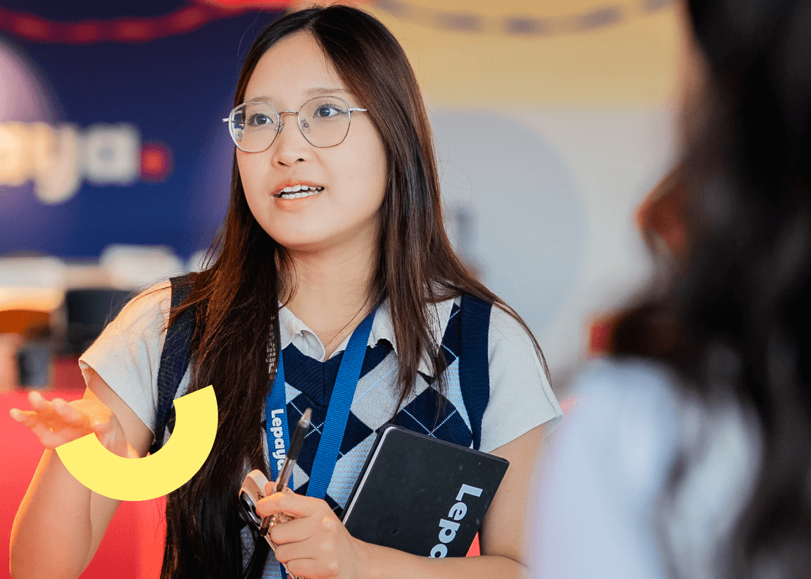 Young woman with glasses and long hair speaking and holding a notebook and pen in an office setting.