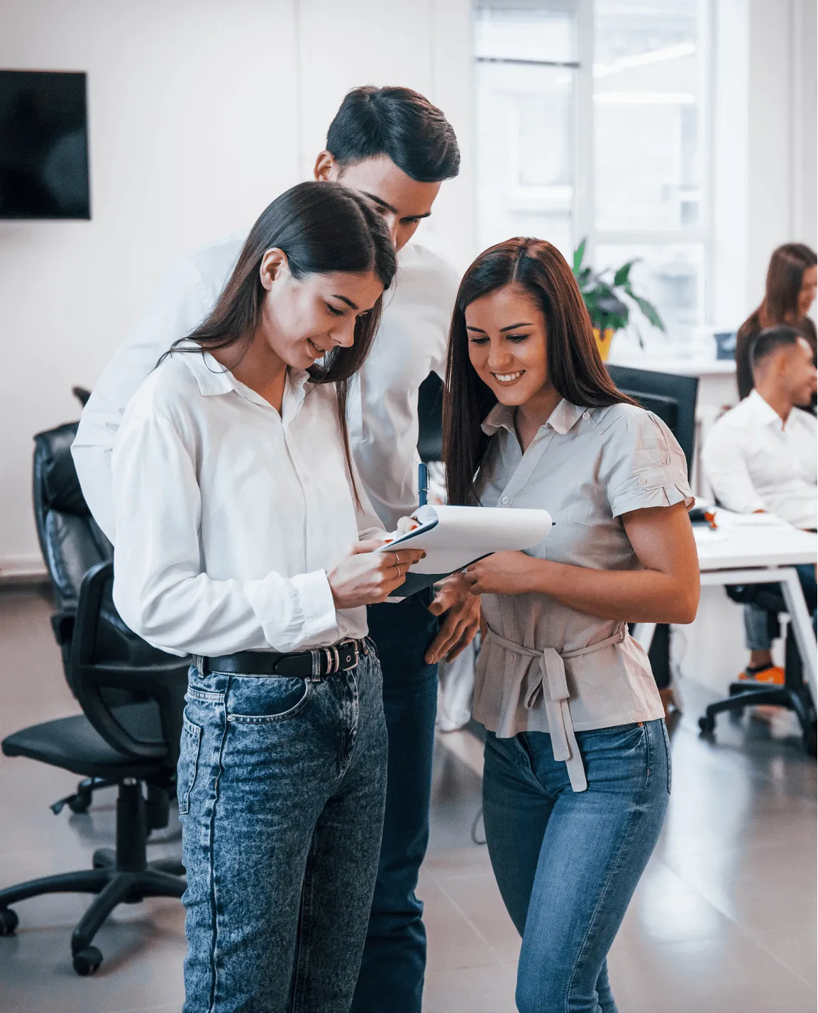 Three young professionals standing together in an office, reviewing notes on a clipboard with smiles.