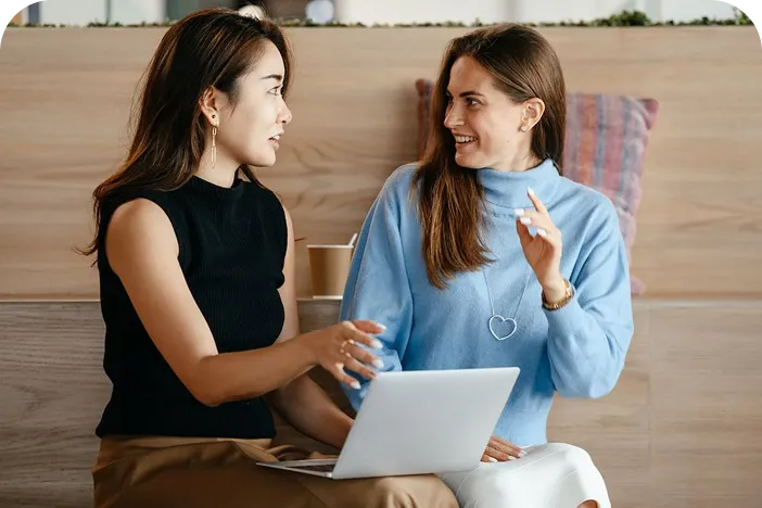 Two women sitting and talking with a laptop, one wearing a black sleeveless top and the other in a blue sweater with a heart necklace.