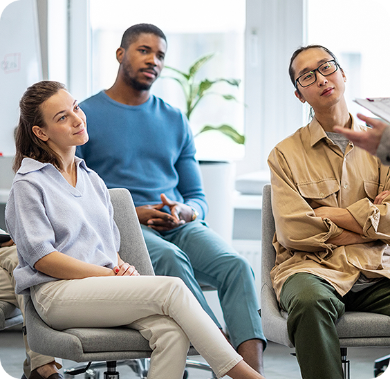 Three diverse adults attentively listening to someone speaking during a casual meeting in a bright office.