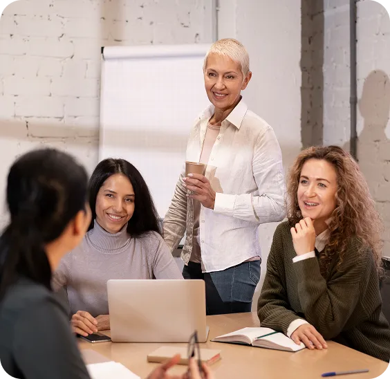 Four women in a casual meeting room engaging in a friendly discussion with a laptop and a notebook on the table.