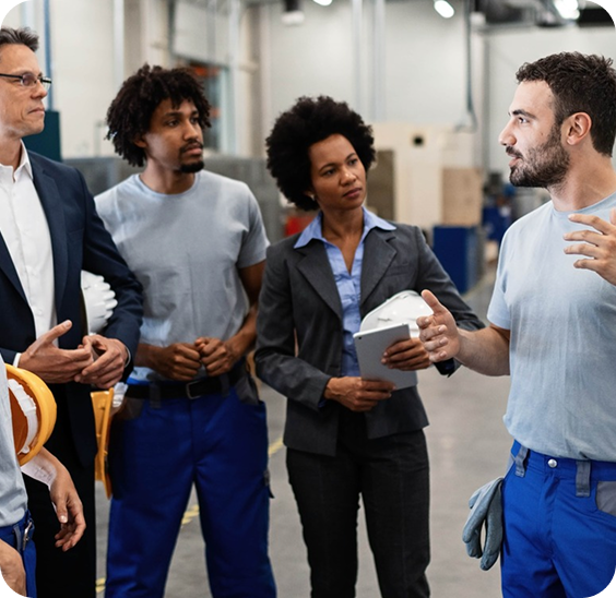 Group of diverse professionals, including two workers in blue pants and a woman in a suit holding a tablet and helmet, listening to a colleague speaking in an industrial setting.