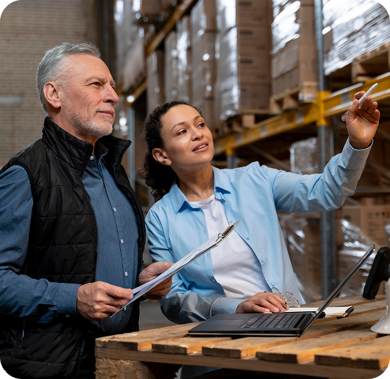 Two warehouse workers inspecting inventory, one holding a clipboard and the other pointing while using a laptop on a wooden pallet.
