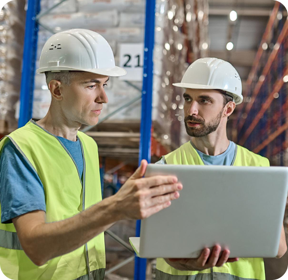 Two warehouse workers wearing white hard hats and yellow safety vests discussing while looking at a laptop in a storage aisle.