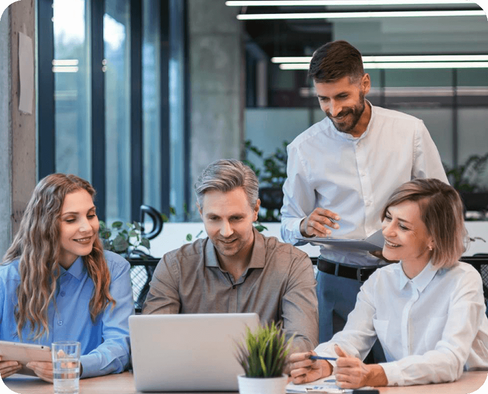 Four colleagues collaborating around a laptop in a modern office with large windows.